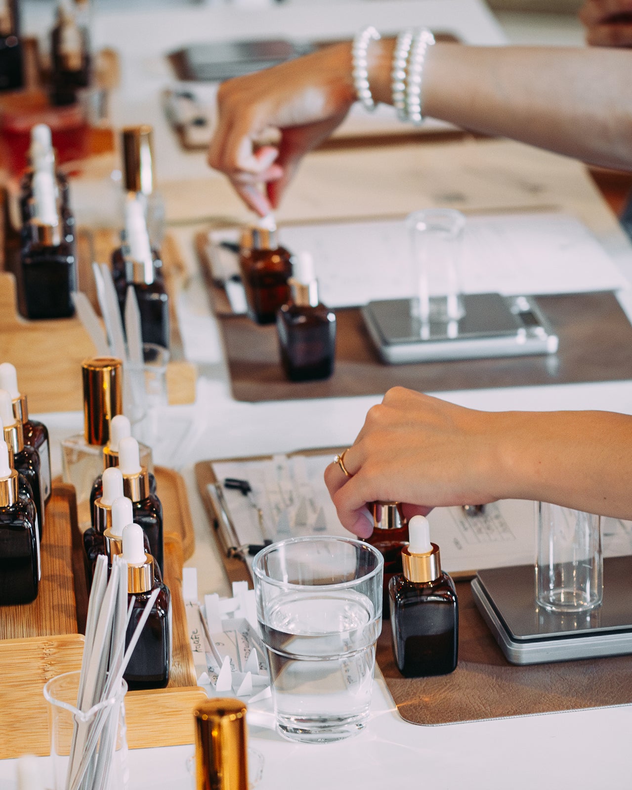 Mâinile a doi participanți lucrând în paralel la atelierul de parfum la masa de lucru cu esențe, blottere și cântare
