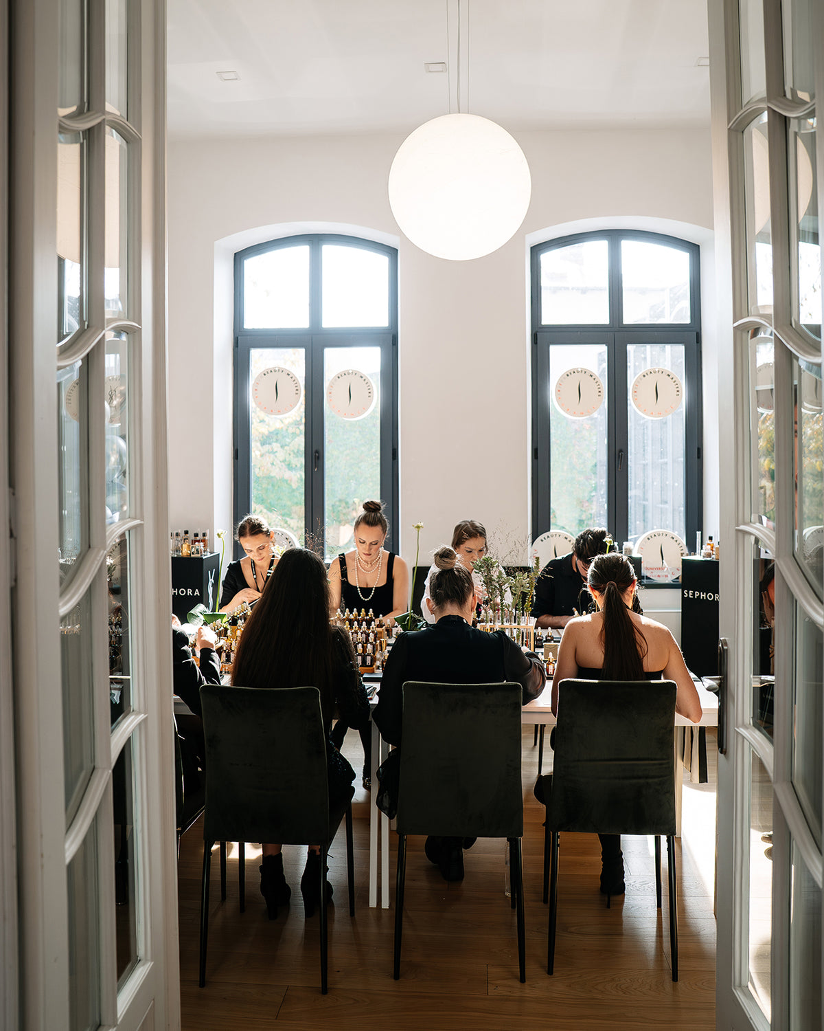 Participanți la un atelier de parfum de grup, lucrând cu esențe la mese, într-o sală cu ferestre înalte
